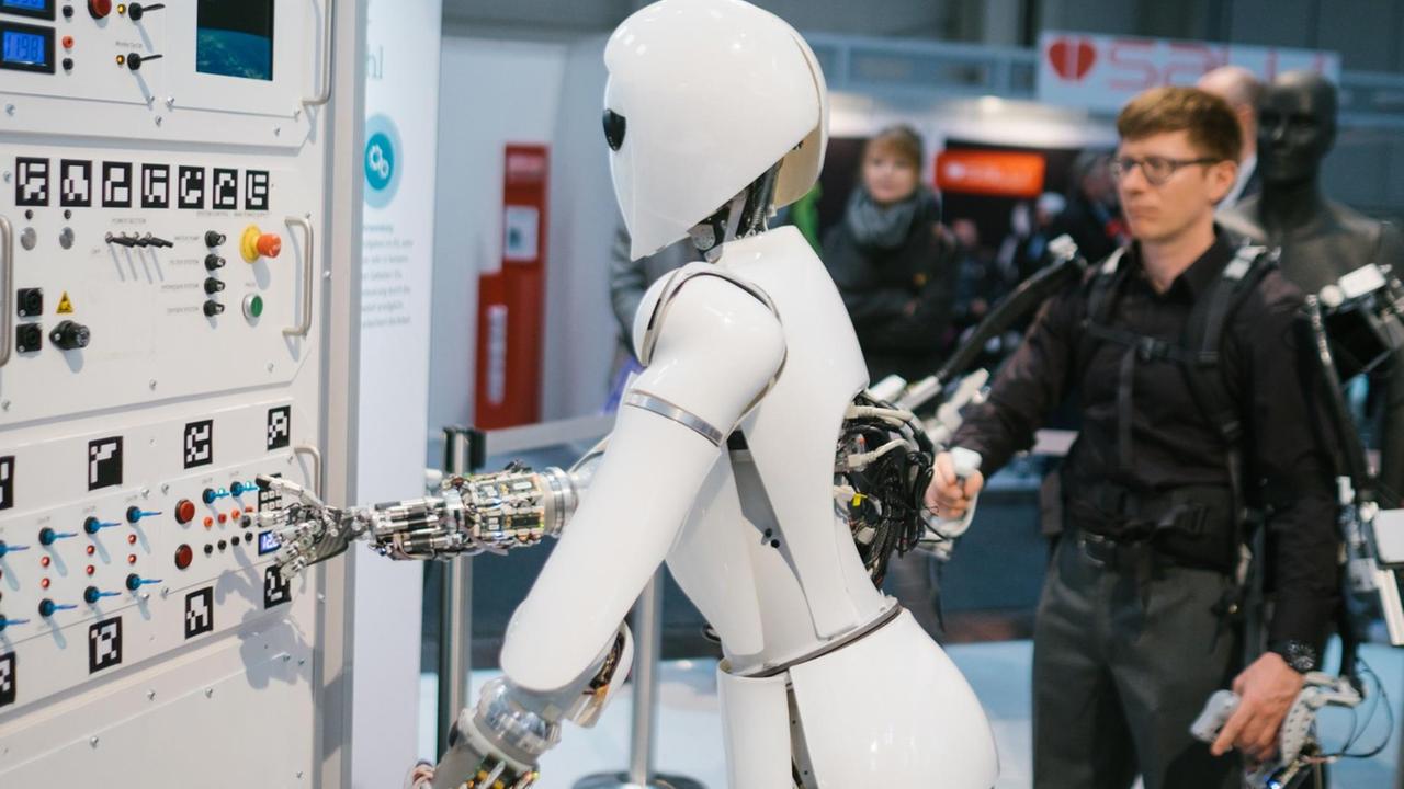 Ein Mitarbeiter des Robotic Innovation Centers steuert am 20.03.2017 bei der CeBIT Messe in Hannover (Niedersachsen) am Stand des deutschen Forschungszentrums für Künstliche Intelligenz (DFKI) mit Hilfe eines Exoskeletts den Roboter AILA.