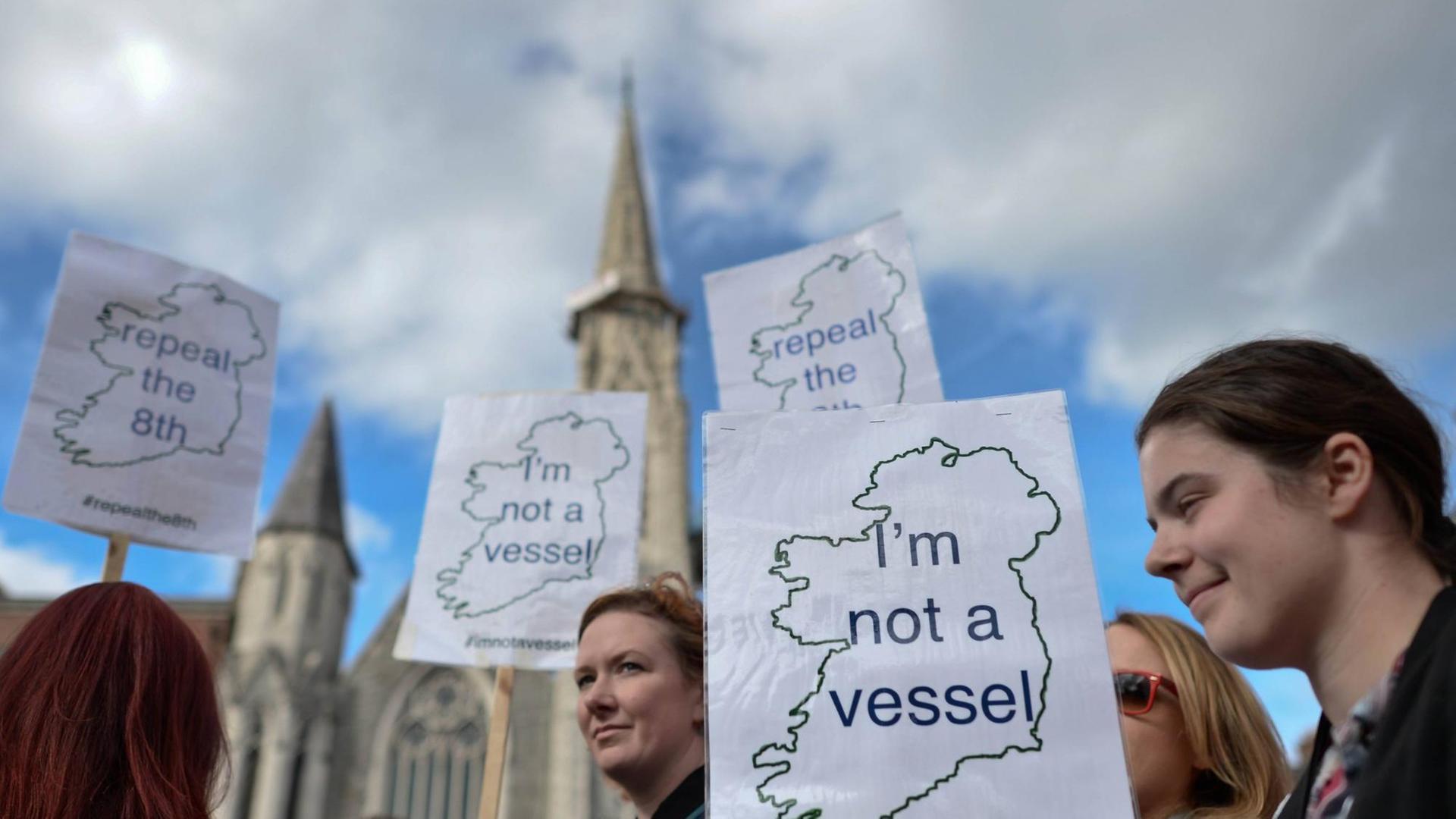 Aktivistinnen demonstrieren mit Schildern, mit der Aufschrift "I'm not a vessel" und den Umrissen von Irland, für die Entkriminalisierung von Schwangerschaftsabbrüchen. Im Hintergrund ist ein Kirchturm zu sehen.