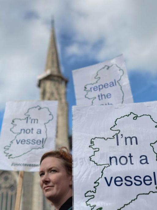 Aktivistinnen demonstrieren mit Schildern, mit der Aufschrift "I'm not a vessel" und den Umrissen von Irland, für die Entkriminalisierung von Schwangerschaftsabbrüchen. Im Hintergrund ist ein Kirchturm zu sehen.