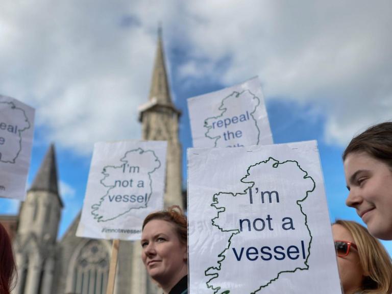 Aktivistinnen demonstrieren mit Schildern, mit der Aufschrift "I'm not a vessel" und den Umrissen von Irland, für die Entkriminalisierung von Schwangerschaftsabbrüchen. Im Hintergrund ist ein Kirchturm zu sehen.