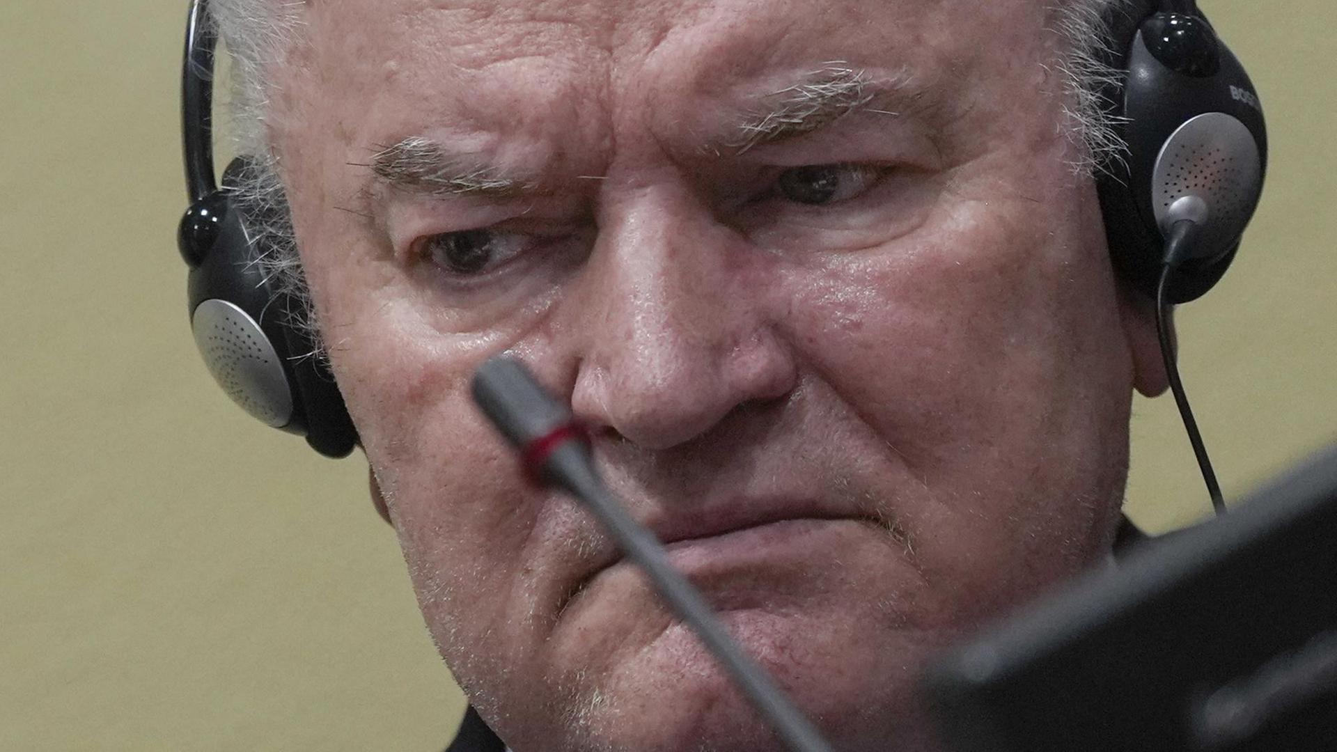 Das Bild zeigt Ratko Mladic mit ernster Mine in einem Gerichtssaal mit einem Kopfhörer und einem Mikrofon.