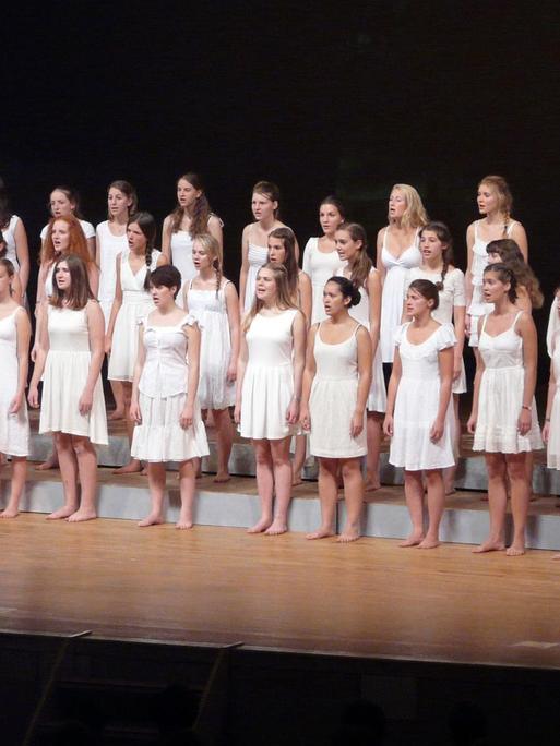 Etwa 40 Mädchen, stehen barfuß in drei Reihen. Sie tragen alle weiße Kleider. Die Chorleitern vorn am Pult trägt ein rotes Kleid.