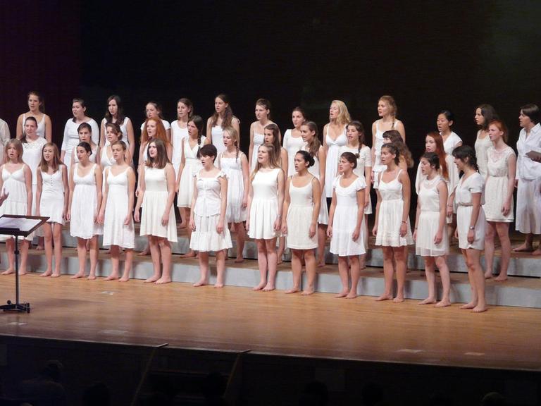 Etwa 40 Mädchen, stehen barfuß in drei Reihen. Sie tragen alle weiße Kleider. Die Chorleitern vorn am Pult trägt ein rotes Kleid.