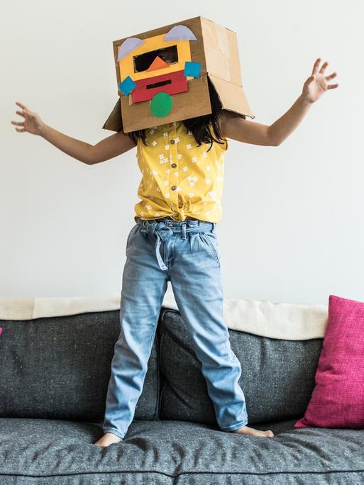 Ein Kind springt auf einem Sofa. Über dem Kopf trägt es einen Pappkarton mit einem bunten Gesicht.