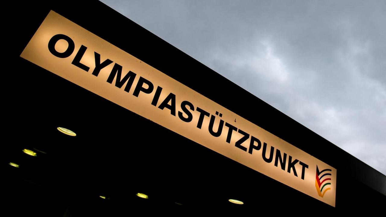 Das Bild zeigt das beleuchtete Schild mit der Aufschrift "Olympiastützpunkt" an der Eingangsdecke von unten fotografiert, am oberen Bildrand sieht man grauen bewölkten Himmel.