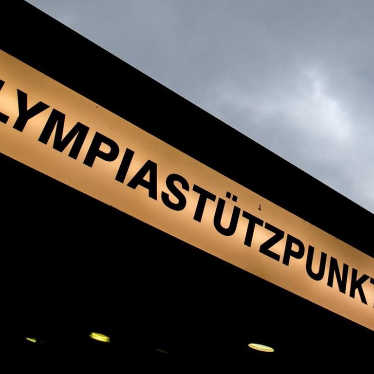 Das Bild zeigt das beleuchtete Schild mit der Aufschrift "Olympiastützpunkt" an der Eingangsdecke von unten fotografiert, am oberen Bildrand sieht man grauen bewölkten Himmel.
