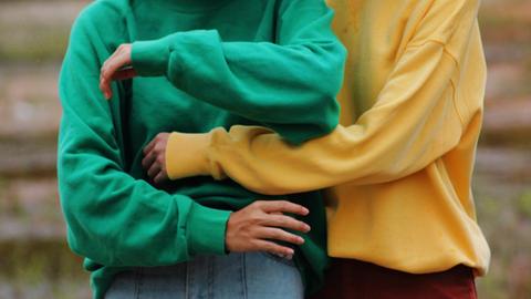 Detail von zwei Oberkörpern. Eine Person in gelbem Pullover umarmt eine Person in grünem Pullover.