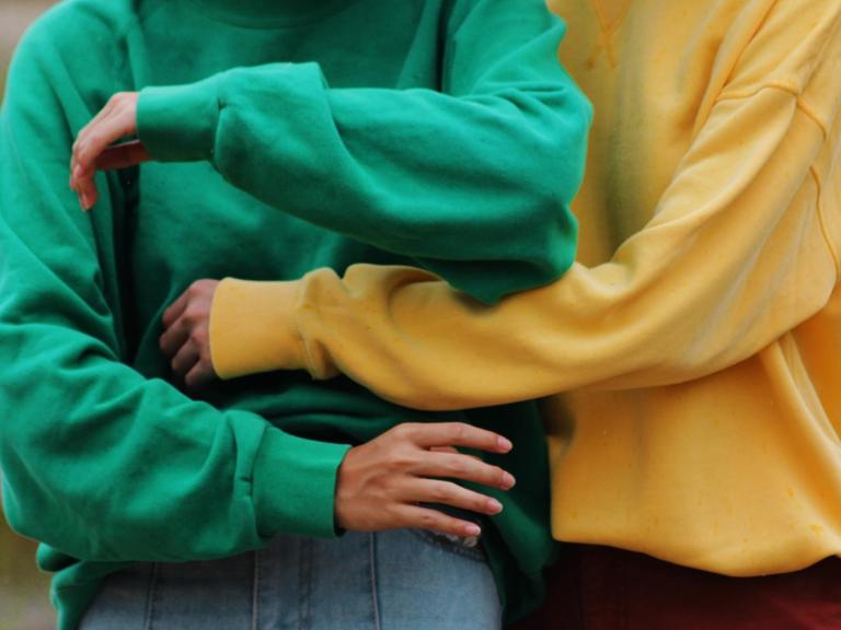 Detail von zwei Oberkörpern. Eine Person in gelbem Pullover umarmt eine Person in grünem Pullover.