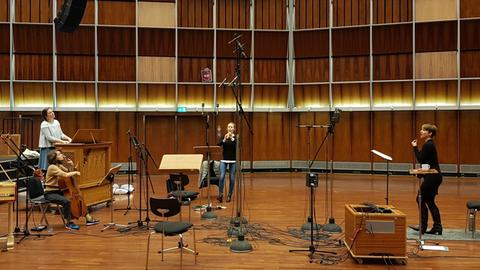 Im holzgetäfelten Deutschlandfunk Kammermusiksaal ist eine ganze Reihe von Standmikrophonen aufgebaut, die Sängerin steht rechts, sie trägt Stiefel und eine blaue Hose und einen blauen Pullover, links steht die Organistin an einer kleinen tragbaren Orgel, davor sitzt die Cellistin, in der Mitte des Bildes steht die Oboistin.