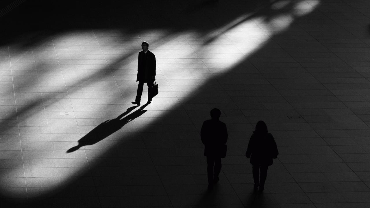 Ein Mann geht auf auf einem Platz, der Schatten vorne. Daneben zwei Menschen, nicht gut erkennbar ausßerhalb des Lichts.