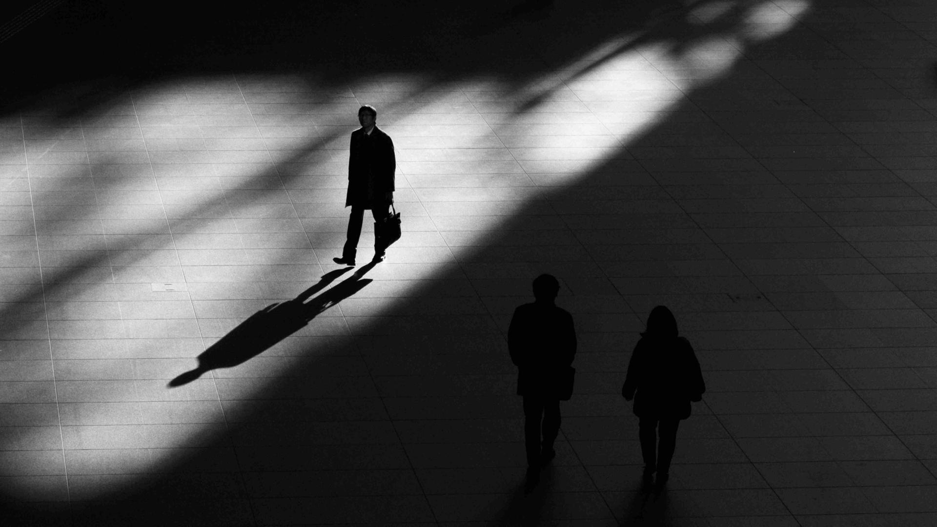 Ein Platz, geteilt in Licht und Schatten: Ein Mann geht im Licht und wirft einen langen Schatten. Auf dem Platz im Schatten zwei Silhouetten, nicht gut erkennbar.