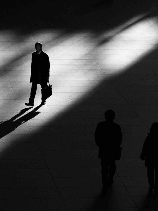 Ein Platz, geteilt in Licht und Schatten: Ein Mann geht im Licht und wirft einen langen Schatten. Auf dem Platz im Schatten zwei Silhouetten, nicht gut erkennbar.