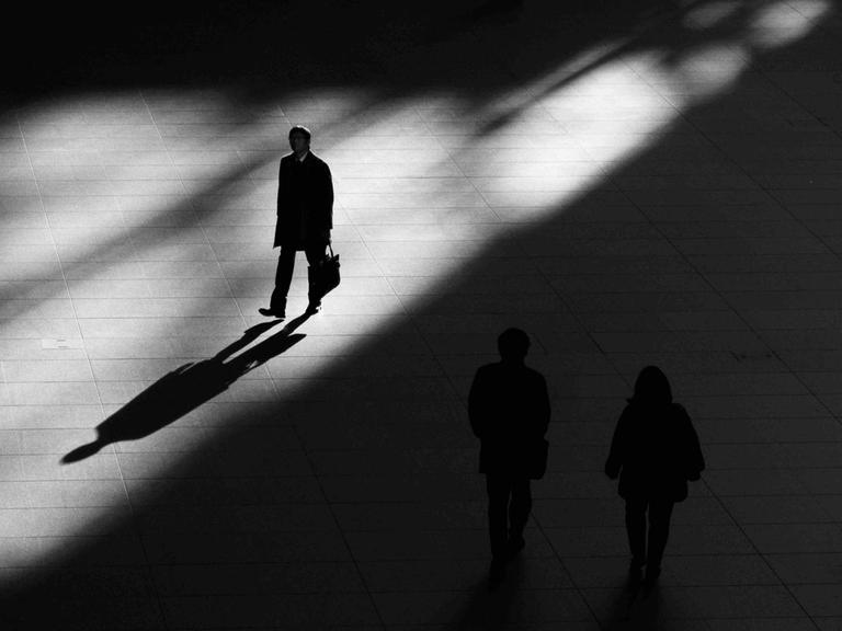 Ein Platz, geteilt in Licht und Schatten: Ein Mann geht im Licht und wirft einen langen Schatten. Auf dem Platz im Schatten zwei Silhouetten, nicht gut erkennbar.