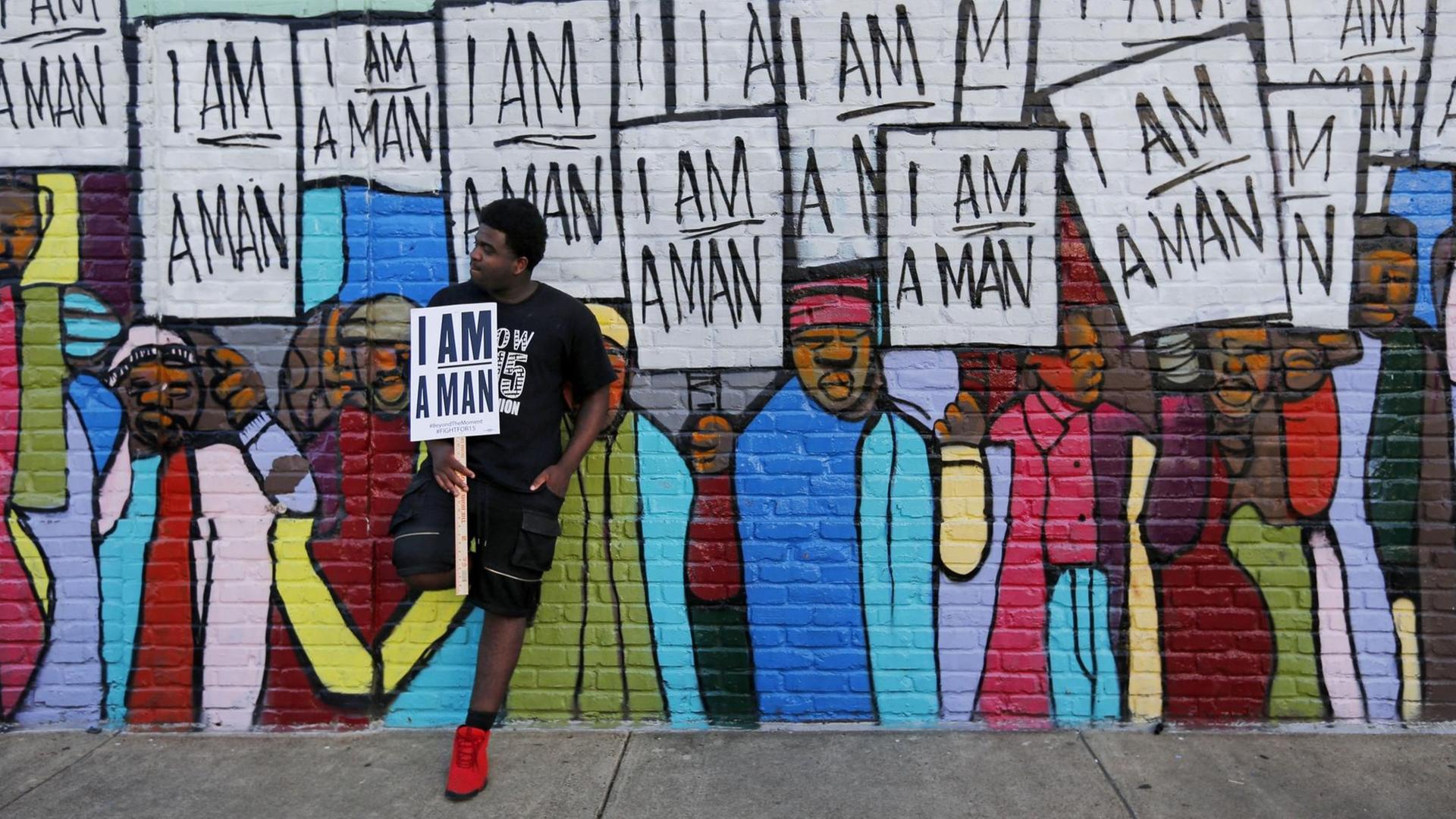 Ein Mann, mit afroamerikanischen Wurzeln, lehnt sich an ein Wandbild mit dem Schild "I am a Man" in der Hand.