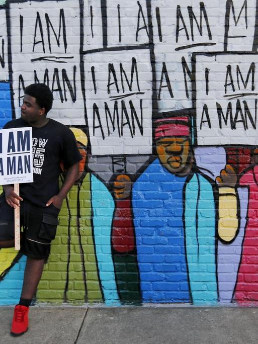 Ein Mann, mit afroamerikanischen Wurzeln, lehnt sich an ein Wandbild mit dem Schild "I am a Man" in der Hand.