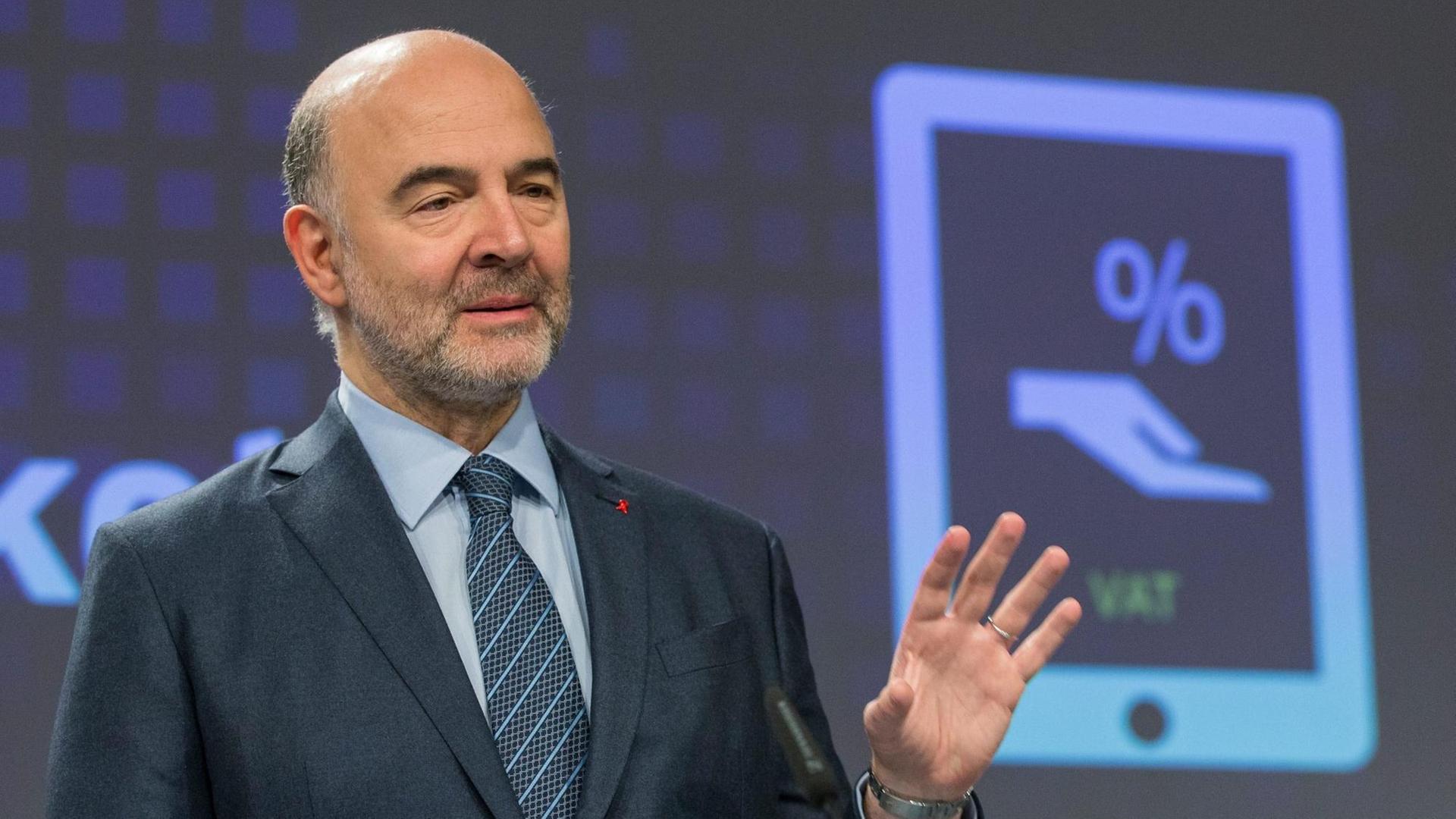 Pierre Moscovici spricht bei einer Pressekonferenz im Dezember 2016 inm Brüssel . Im hintergrund ist ein stilisiertes Tablet abgebildet, auf dem eine stilisierte Hand und ein prozentzeichen zu sehen sind.
