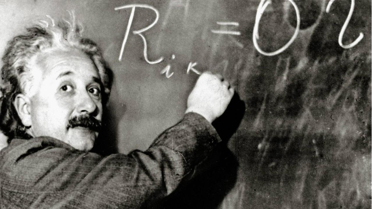 Albert Einstein schreibt eine Formel an die Tafel und guckt dabei in die Kamera.