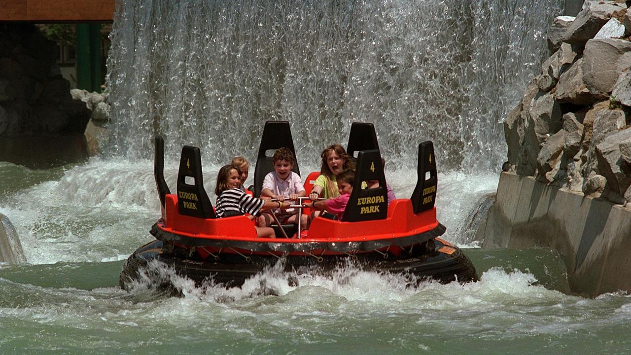 Das Bild zeigt Besucher auf einem Schlauchboot. Sie fahren durch einen Wildwasserkanal. Neben dem Kanal sind Felsen zu erkennen und im Hintergrund ein Wasserfall.