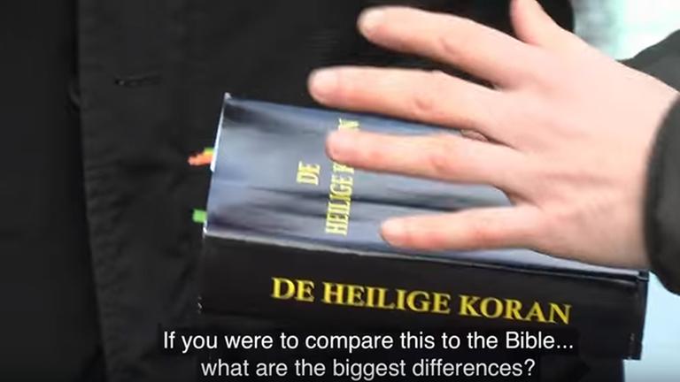 Eine Hand hält ein Buch mit dem Umschlag "De Heilige Koran", der Untertitel besagt: "If you were to compare this to the Bible... what are the biggest differences?"