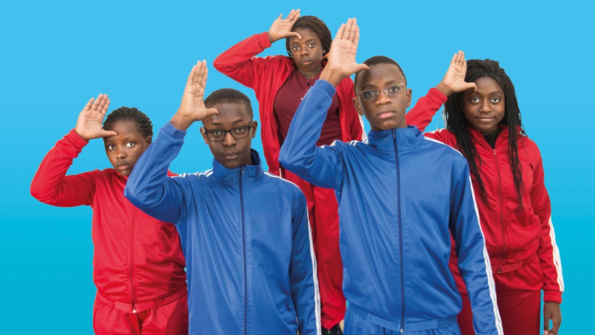 Fünf dunkelhäutige Kinder in roten und blauen Trainingsanzügen stehen in einer Gruppe zusammen und machen den DDR-Pioniergruß mit einer Hand an der Stirn.