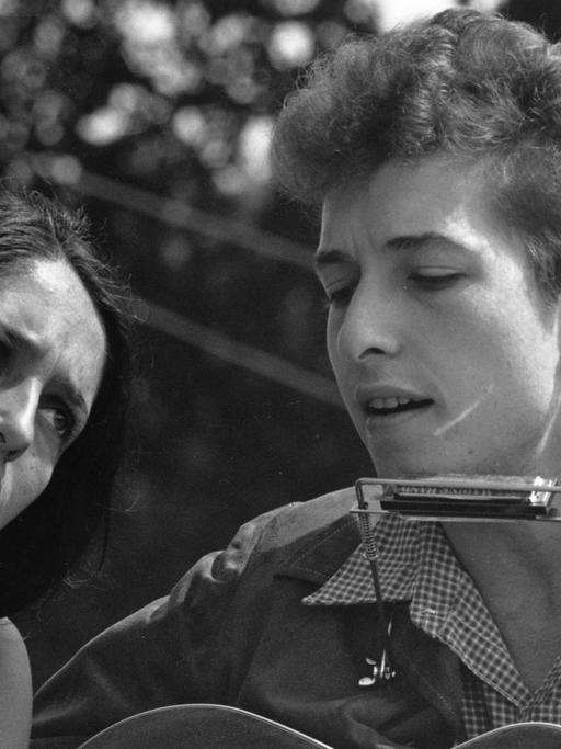 Ein Schwarzweiß-Foto zeigt eine Frau in Sommerkleid gelehnt an die Schulter eines Mannes mit Akustik-Gitarre und Mundharmonika Das Traumpaar des Folk: Joan Baez und Bob Dylan Hier 1963 am Rande des "Marschs auf Washington "1963