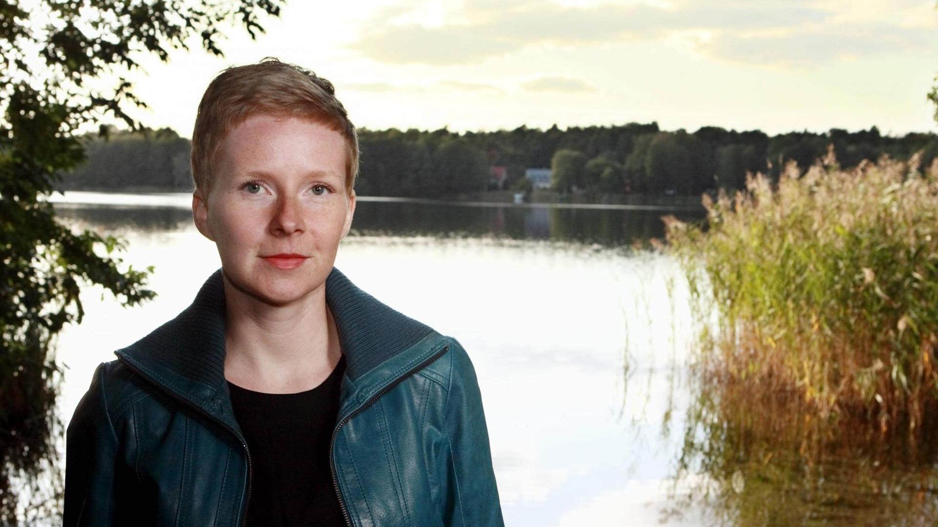 Die Autorin Judith Zander sitzt vor einem See und schaut in die Kamera.