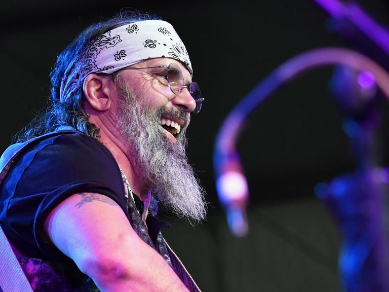 Steve Earle mit Stirnband und Gitarre steht auf einer Bühne und lacht ins Publikum.