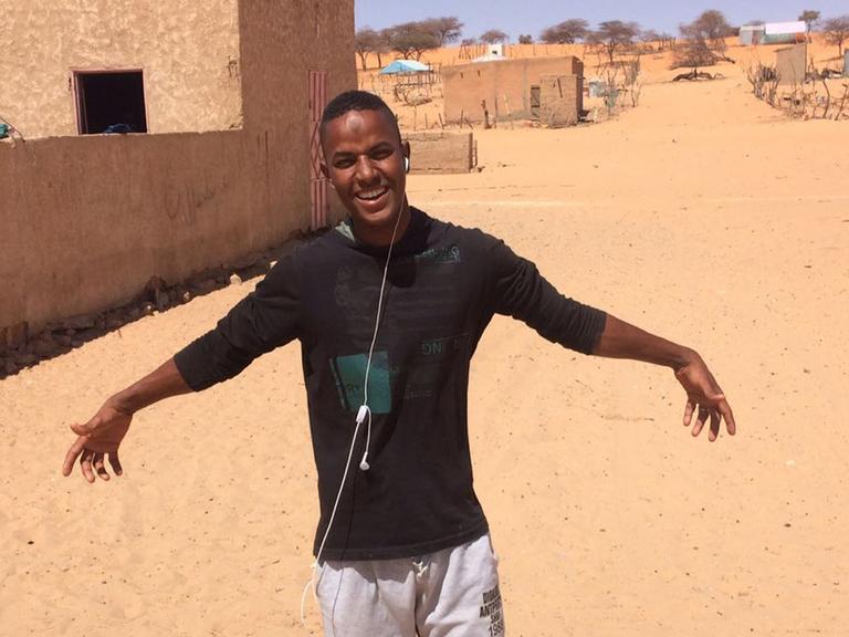 Ein junger Mann steht in einem Dorf auf einem gelben Sandboden, hat die Arme ausgebreitet und lächelt in die Kamera.