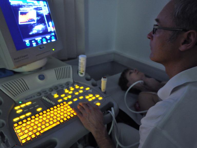 Ein Arzt sitzt in einem dunklen Raum vor einem Bildschirm eines Ultraschallgeräts, in der rechten Hand ein Ultraschallgerät, das an der Halsschlagader einer Patientin liegt.