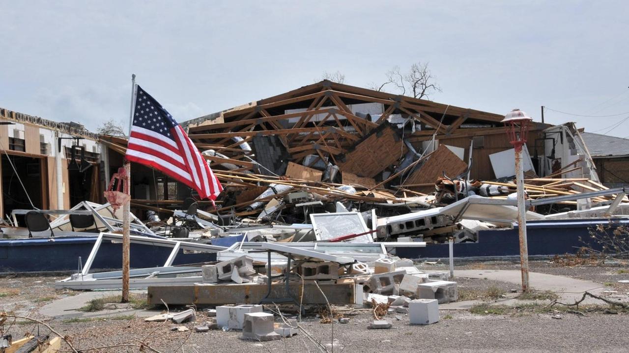 Man sieht ein zerstörtes Haus, davor weht eine US-Flagge