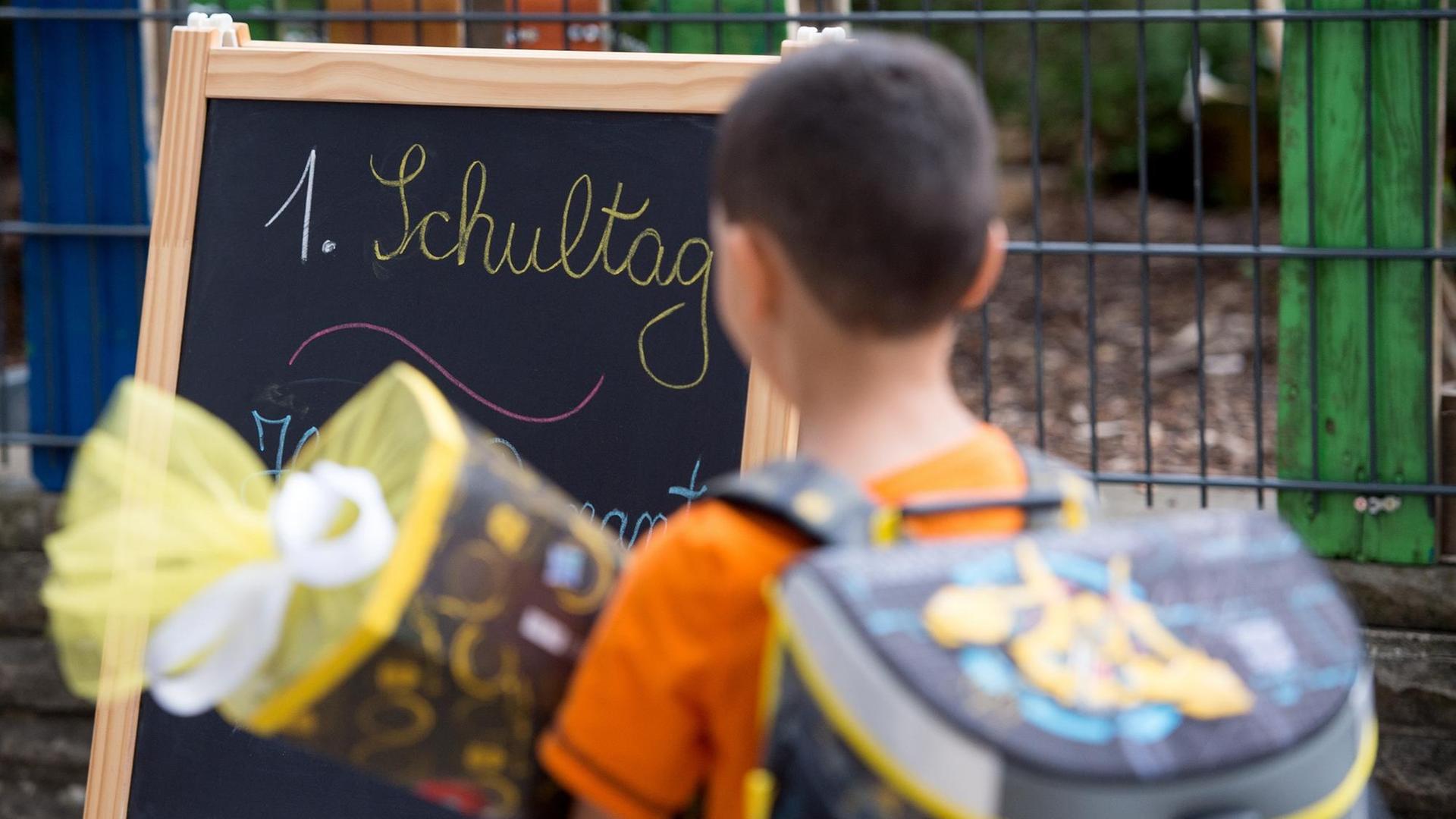 Ein Junge steht mit Schul-Tüte und Ranzen vor einer Tafel. Darauf steht: 1. Schul-Tag.