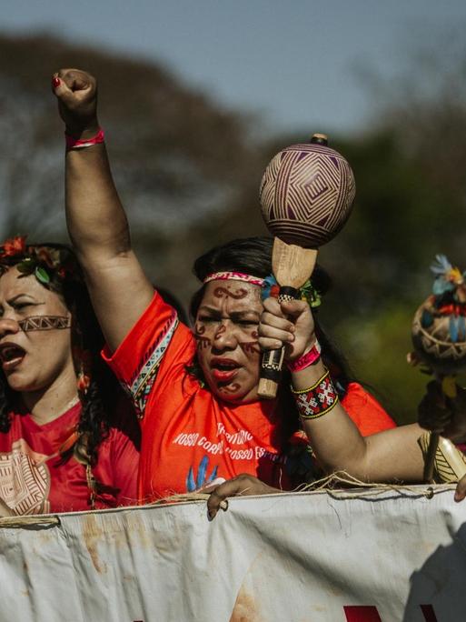 Fünf indigene Frauen protestieren in traditioneller Kleidung und Kopfschmuck. Sie strecken die Arme und teilweise die Fäuste nach oben. Am Rand steht ein Polizist, der sie beobachtet.