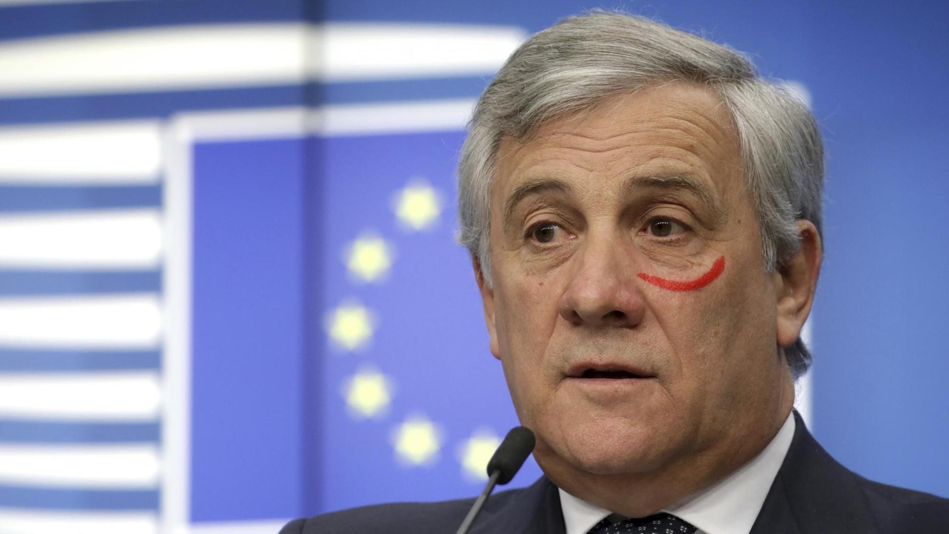Tajani hinter einem Mikrofon. Unter dem rechten Auge trägt er einen roten halbkreisförmigen Strich.