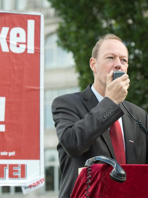 Martin Sonneborn spricht durch ein Megaphon, neben ihm ein Wahlplakat mit dem Slogan "Merkel ist doof!"