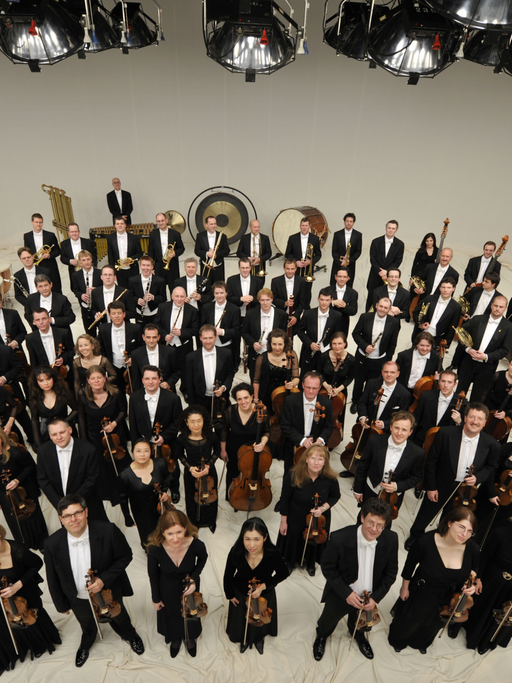 Die Mitglieder des Orchesters stehen mit ihren Instrumenten in einem großen Raum und schauen nach oben in die Kamera.
