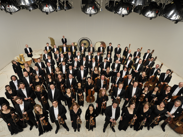 Die Mitglieder des Orchesters stehen mit ihren Instrumenten in einem großen Raum und schauen nach oben in die Kamera.