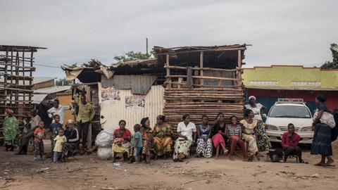 Frauen in afrikanischer Kleidung sitzen vor einem Unterstand in einer Siedlung. Daneben steht ein weißes Auto.