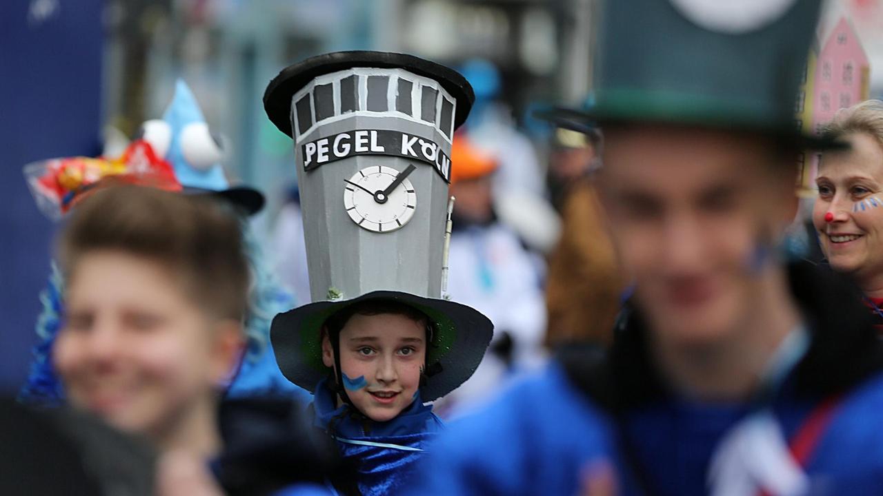 Ein Junge geht in Köln bei den "Schull- un Veedelszöch" mit dem Kölner Pegelhaus auf dem Kopf über die Straße.
