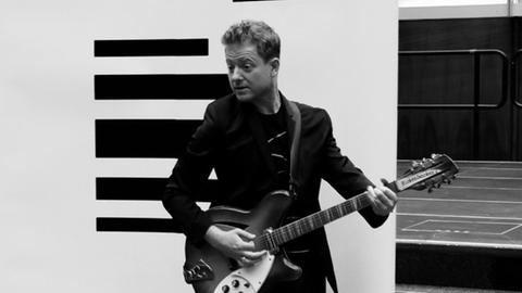 Schwarz-Weiß-Aufnahme: Musikkabarettist Martin Zingsheim steht vor einem DLF-Banner und spielt scheinbar auf eine E-Gitarre.