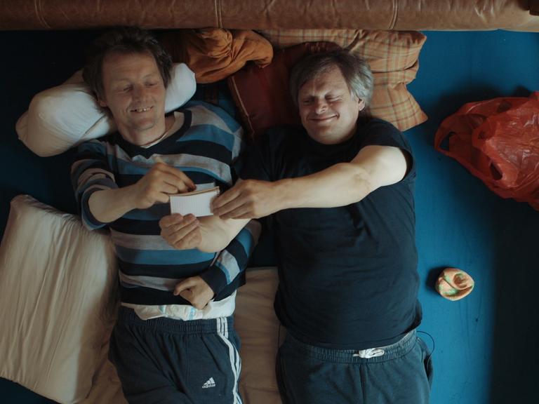 Filmszene aus Bruderliebe: Markus und Michael liegen nebeneinander auf dem Teppich und schauen sich eine Seite in einem kleinen Block an.