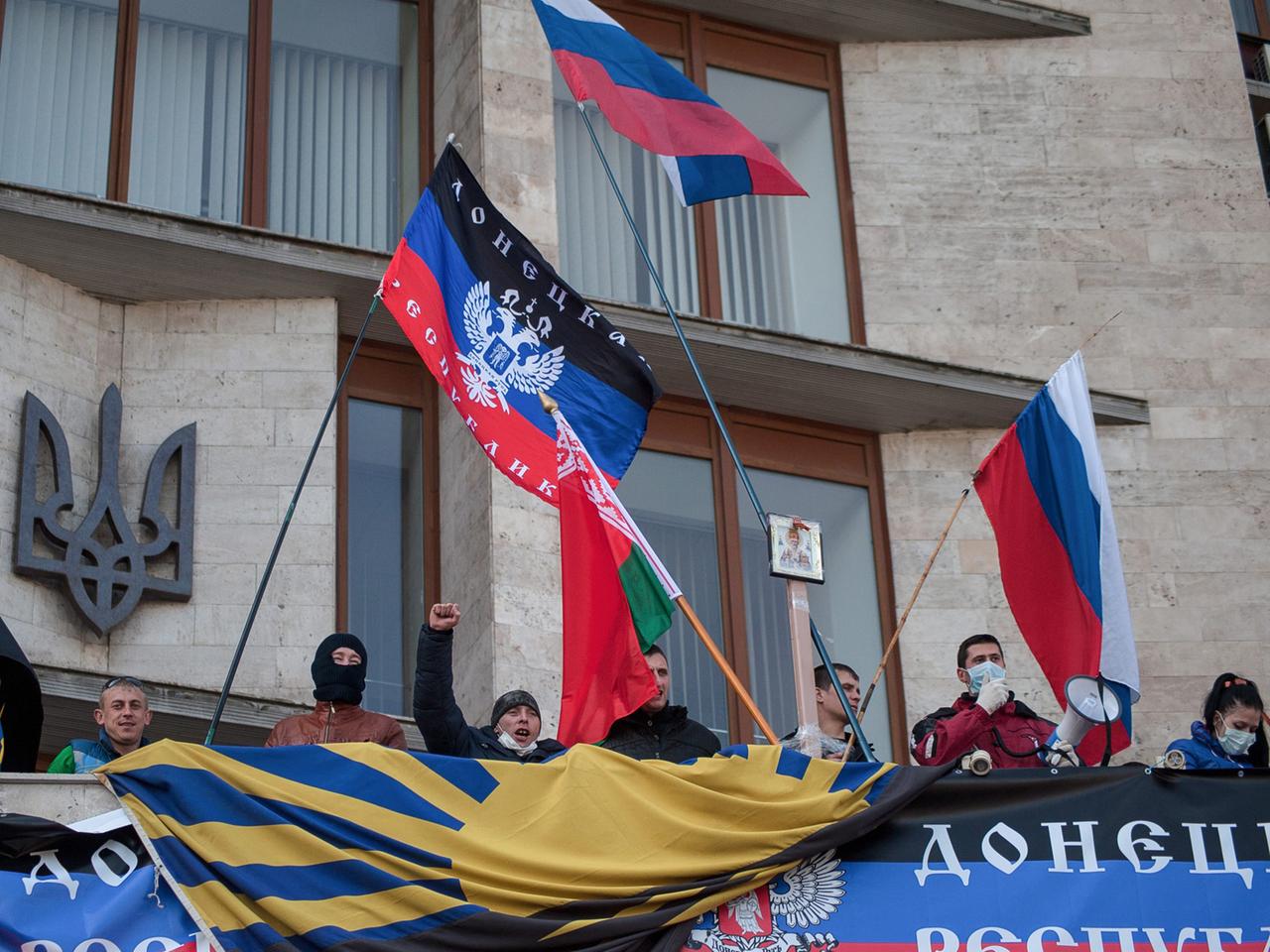 Vor einem Verwaltungsgebäude sind teils vermummte Demonstranten zu sehen, die die russische Flagge schwenken und skandieren.