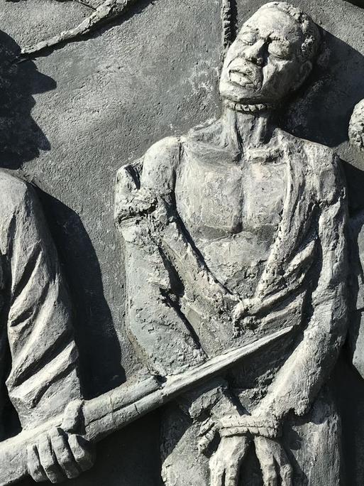 Denkmal in Windhoek für den von deutschen Kolonialtruppen begangenen Völkermord an den Herero und Nama: An einem Ast hängen die an den Händen gefesselten Leichen eines Mannes und einer Frau. Daneben steht ein kräftiger Mann mit einem Gewehr.