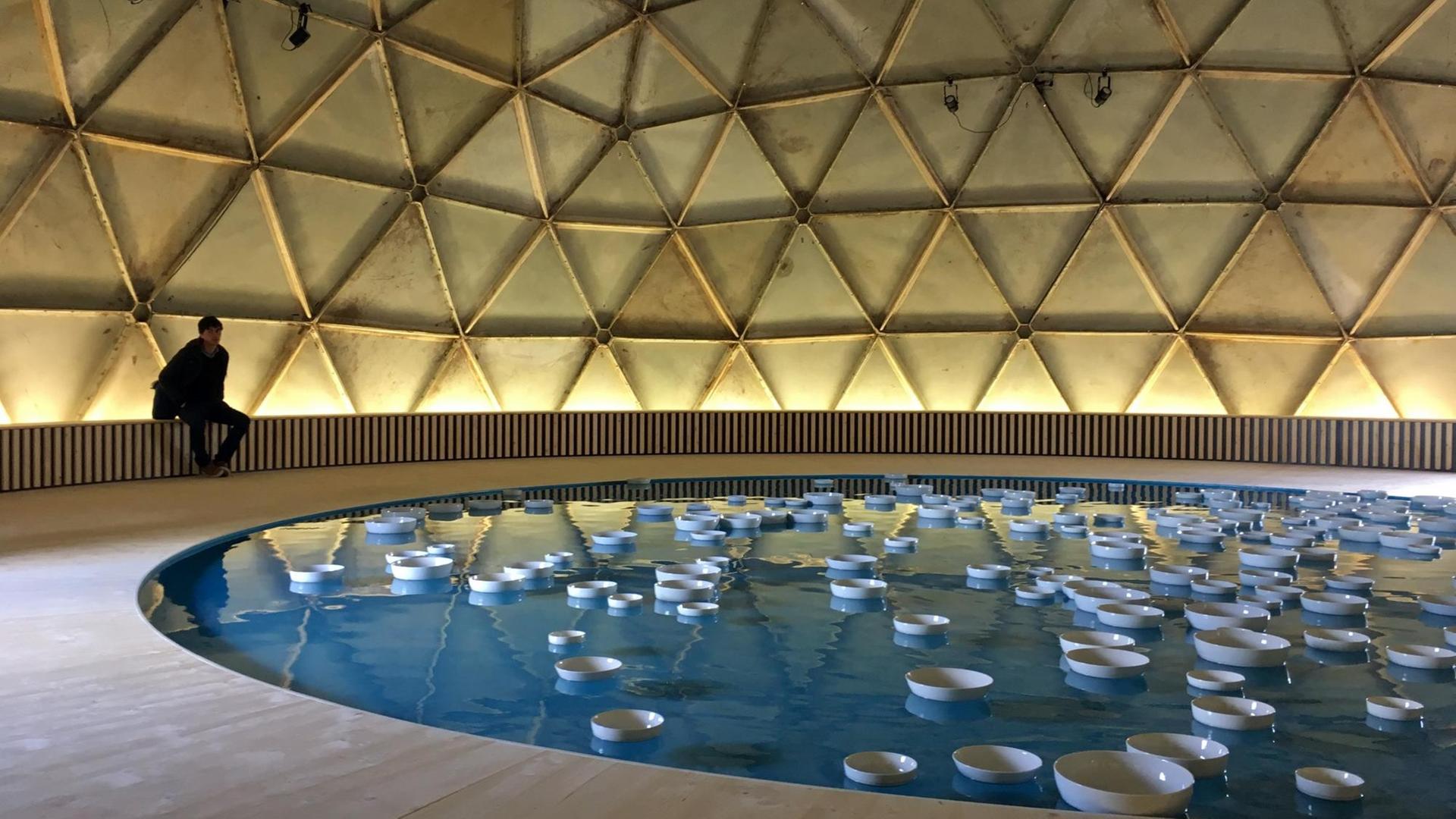 Ein Werk des 1983 gestorbenen amerikanischen Künstlers Richard Buckminster: In einem runden Wasserbecken, schwimmen um die 40 weiße Keramikschalen. Durch die sanften Wellenbewegungen des Wassers stoßen sie immer wieder aneinander.