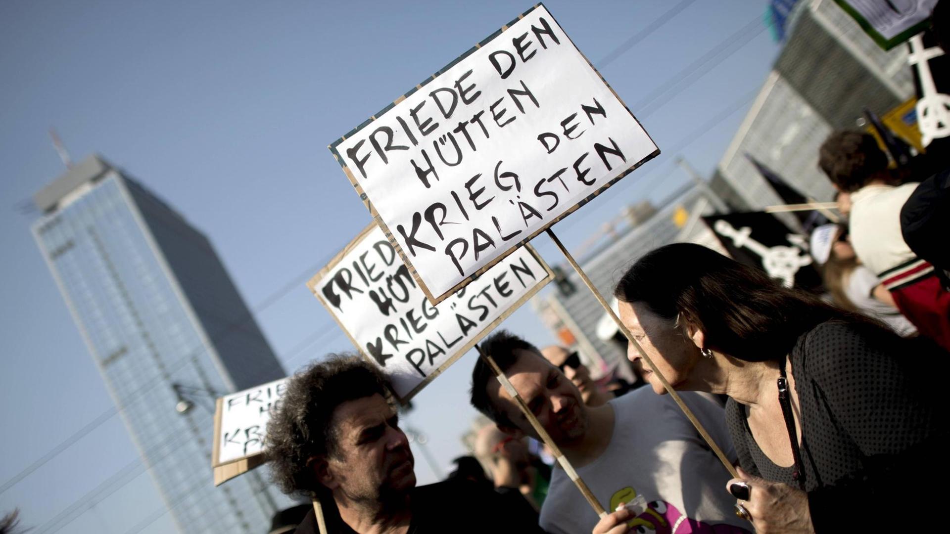 Drei Menschen stehen bei der Mietendemo in Berlin zusammen und halten ein Schilder mit der Aufschrift "Friede Den Hütten - Krieg Den Palästen".