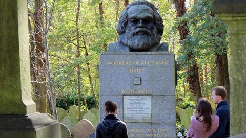Das Bild zeigt den massiven Grabstein mit der Marmorplatte und der Marx-Büste darauf von vorne. Dahinter Bäume, vor dem Grab drei Menschen.