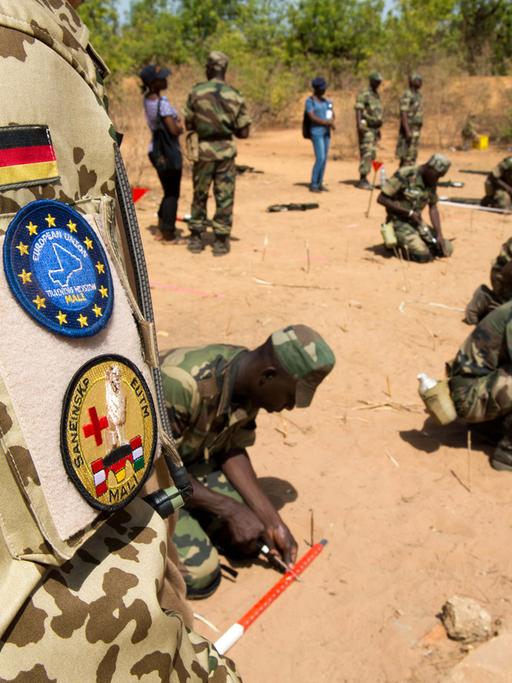 Die Schulter eines Bundeswehrsoldaten in Uniform ist zu sehen, dahinter knien Pioniere der Armee Malis.