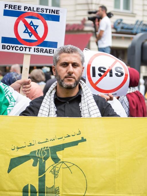 Ein Demonstrationsteilnehmer haelt die gelb-gruene Fahne der islamistischen Hisbollah-Partei und seine Mitdemonstranten haben Schilder gegen die Terrororganisation Islamischer Staat, IS.