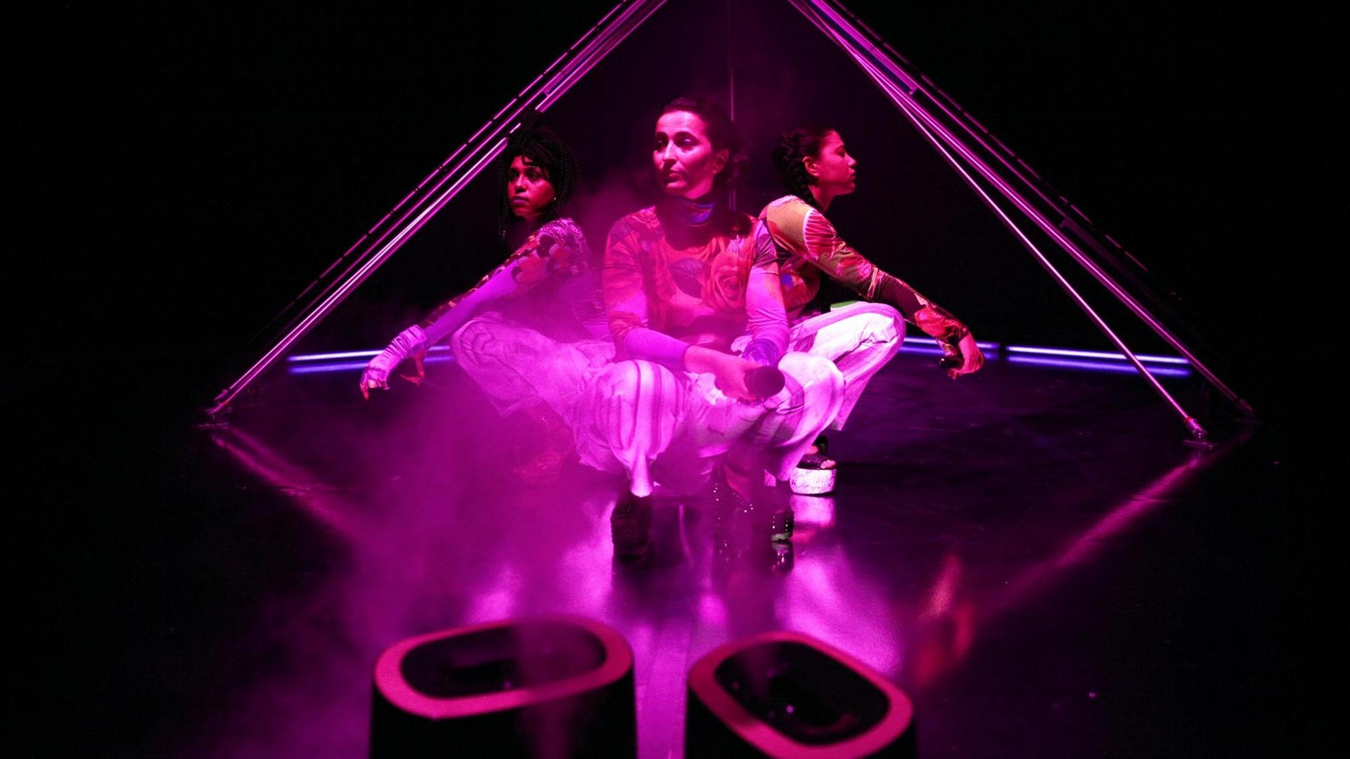 Drei Frauen hocken Rücken an Rücken auf einer Bühne, pink angestrahlt.
