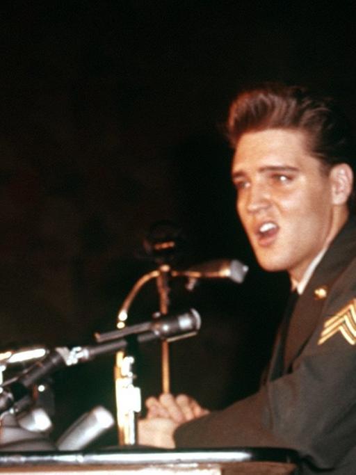 Elvis Presley sitzt an einem Tisch, auf dem Mikrofone stehen.