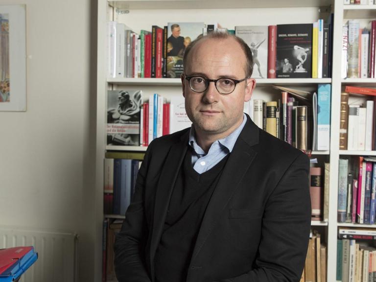 Johann Chapoutot vor einer Bücherwand. Er hat eine Brille auf und trägt ein helles Hemd und einen dunklen Pullover